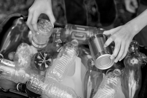 A close-up black-and-white photograph showing two hands sorting recyclable waste among a pile of various plastic bottles and containers. One hand holds a metal can, while the other hand picks up a plastic bottle. The plastic bottles vary in shape, size, and texture, with some appearing transparent and others slightly opaque. The scene appears to be outdoors, possibly on a paved surface or driveway, with the edge of the waste pile visible in the background. The focus is on the manual sorting process, highlighting the importance of proper rubbish separation in waste management. This image is relevant to rubbish removal services such as those offered by Garden Clearance Knightsbridge, serving areas including Knightsbridge and surrounding postcode districts, emphasizing environmentally conscious disposal practices.