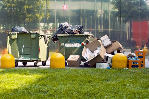A row of three large waste bins situated on a paved area outside a building with glass windows reflecting trees and surroundings. The left bin is green with a closed lid, while the middle bin is also green and partially covered with a black trash bag. The right bin is overflowing with assorted cardboard boxes, some flattened and others still in their original shape, along with plastic containers and packaging materials. Between the bins and on the ground are additional cardboard boxes and plastic waste, some stacked or leaning against the bins. A yellow barrier is positioned in front of each bin to prevent vehicle intrusion. The foreground features a well-maintained grass lawn, and the background shows the reflective glass façade of a modern building, indicating an urban setting that could relate to rubbish removal services in Knightsbridge or nearby areas.