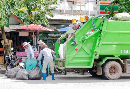 A bright green waste collection vehicle is parked on a street, with its rear hatch open and waste disposal opening visible. Two workers wearing white protective suits, gloves, and boots are actively loading large black rubbish bags into the vehicle’s rear compartment. In the background, there are small market stalls with colorful umbrellas and canopies, some trees providing partial shade, and a mix of buildings. The scene takes place outdoors on a pavement, with a residential or commercial area setting, and appears to be part of a municipal rubbish removal service in an urban environment. The overall atmosphere is utilitarian, focusing on waste collection in a busy area, with visible street activity and nearby street furniture. The image exemplifies rubbish removal services typical in a London neighbourhood, aligning with Garden Clearance Knightsbridge’s offerings for waste collection and clearance tasks in the Knightsbridge area, inferred from the context and surrounding environment.