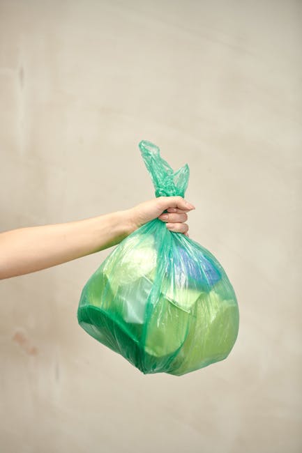 A human hand holds a green and blue plastic bag filled with trash, including visible crumpled paper and discarded materials, against a plain, beige background. The bag is twisted and knotted at the to
