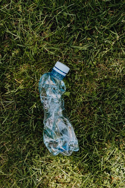A human hand holds a green and blue plastic bag filled with trash, including visible crumpled paper and discarded materials, against a plain, beige background. The bag is twisted and knotted at the to